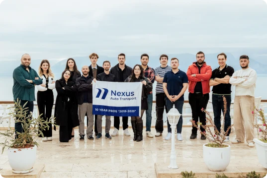 Nexus Auto Transport team holding a 10 Year Pros 2015-2025 anniversary banner at an outdoor seaside gathering